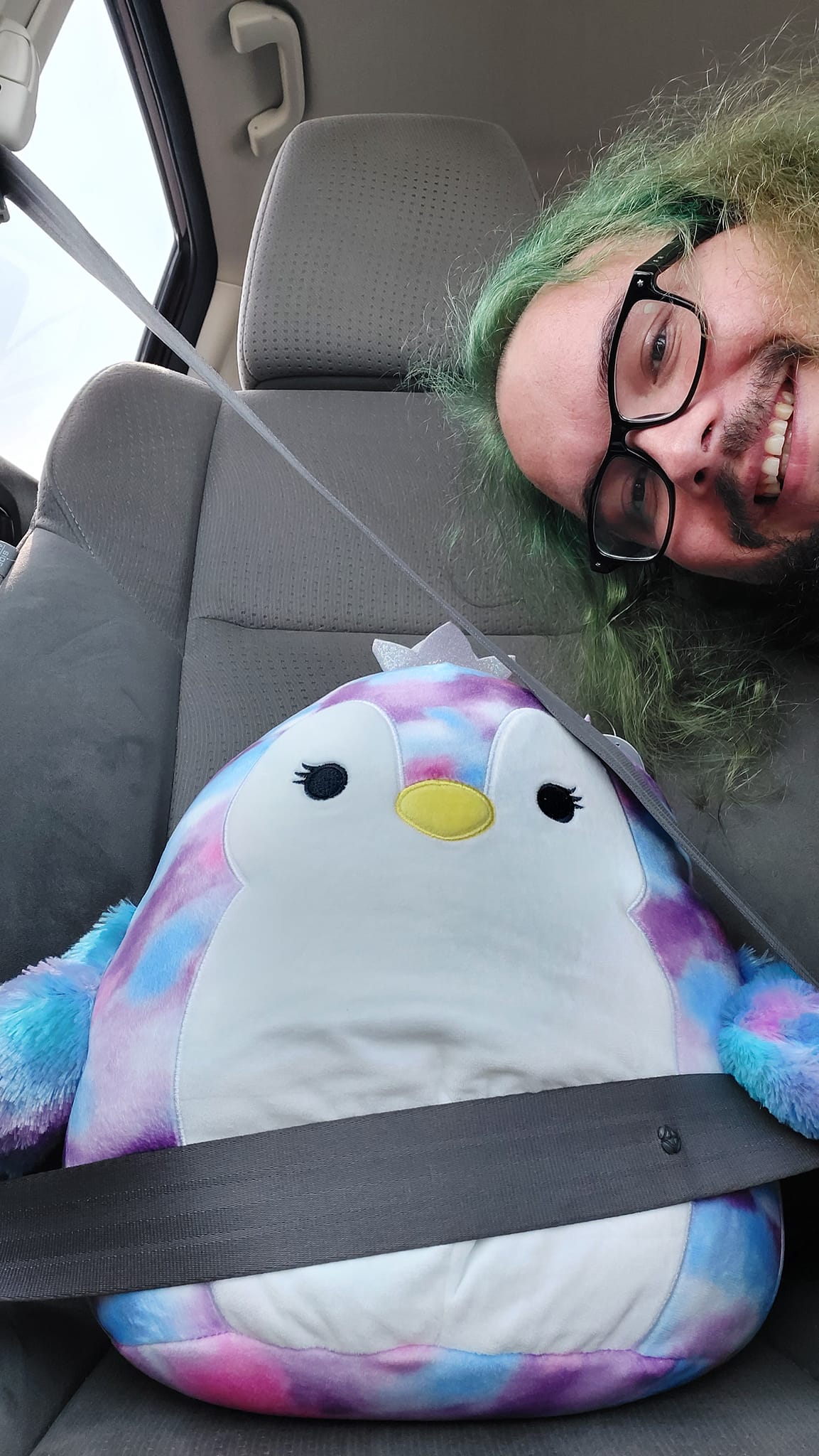 A man with long green hair and a beard pokes his head into frame from the side, the photo centres on a blue, red and purple Squishmallow penguin